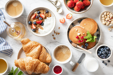 Breakfast table with granola, fresh berries, croissants, pancakes, honey, nuts and coffee on a marble background