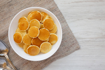 Homemade Mini Pancakes Cereal in a white bowl on a white wooden surface, top view. Flat lay, overhead, from above. Copy space.