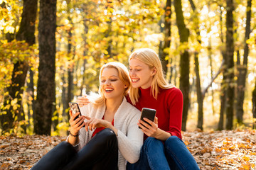 Two excited young girls using mobile phones while sitting in autumn park outdoors and pointing finger.