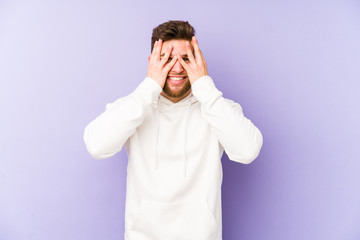 Young caucasian man isolated on purple background blink at the camera through fingers, embarrassed covering face.