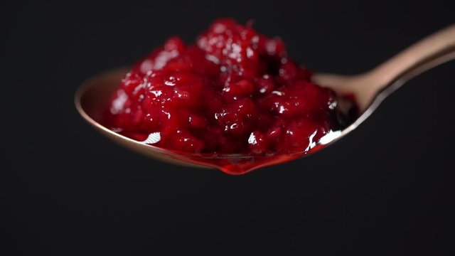 Spoon with red raspberry jam and one drop. Red raspberry jam dripping and flowing down from the metal spoon on black background, close up. Brass spoon with homemade red raspberry jam