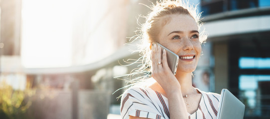 Red haired businesswoman with freckles discussing on phone and posing with a laptop outside in a sunny day