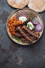 Serbian traditional grilled cevapi sausages and pljeskavicas served with kajmak, prebranac and pitas. Vertical shot on a brown stone background, top view, copy space