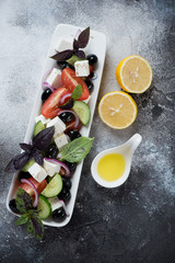 White plate with greek salad, lemon and olive oil over dark grey stone background, vertical shot, top view