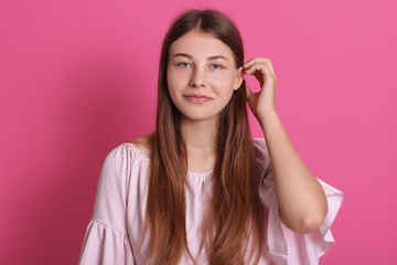 Portrait of young happy woman looks at camera and touching her hair, standing against pink wall, has pleasant appearance, wearing pale pink outfit.