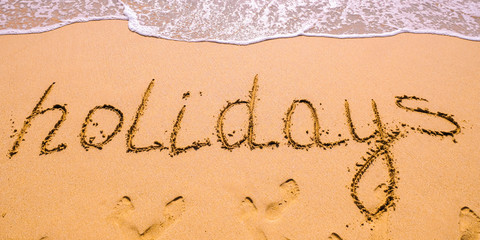 Word Holidays hand written in the sand with a sea wave. Close up sand texture on beach in summer.