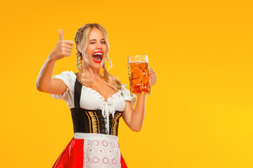 Young sexy Oktoberfest girl waitress, wearing a traditional Bavarian or german dirndl. Woman serving big beer mugs with drink isolated on yellow background.