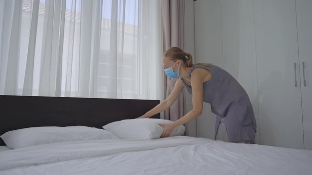 A young woman in a medical face mask changes bed linen in a hotel room. The hotel works as a special quarantine facility for people arriving from abroad. Travel during COVID concept