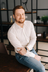 Fototapeta premium Smiling young business man wearing stylish clothing looking at camera while sitting indoors on the chair in modern office. Portrait of bearded handsome gentleman wearing white shirt.
