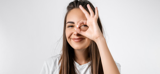 Fototapeta premium Close up portrait of carefree modern girl with long chestnut hairs, looking at the camera through fingers in OKAY gesture. Face expressions, emotions, and body language