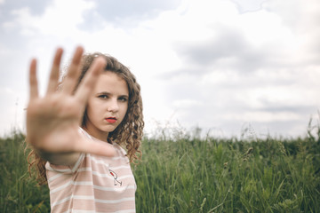 A Young Girl Standing On A Field And Show Stop Hand. Cloudy Aay, Green Meadow. Blurred Background.