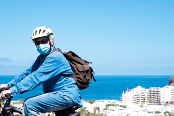 Retired senior man in outdoor excursion with bicycle and yellow helmet wearing face mask due to coronavirus. Blue sky and horizon over the sea.