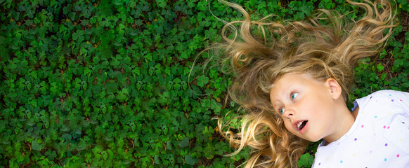 Child girl with blond hair on green grass background. A child with a smile lies on the grass in the...
