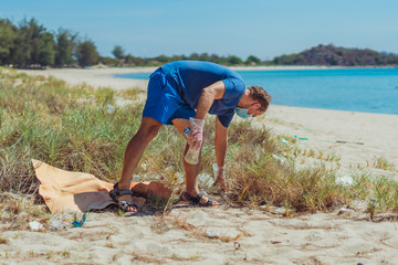 Volunteer in blue face mask lazur sea sand beach. Active man pick up garbage plastic foam bags...