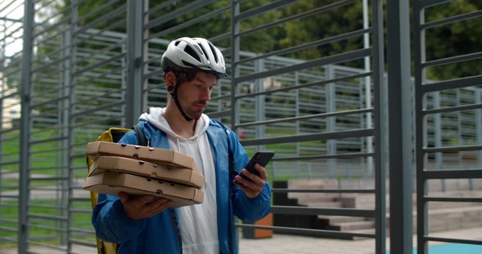 Male person in protective cycling helmet with bag carrying pizza boxes at street. Man courier delivering order to customer while using smartphone. Concept of delivery food services