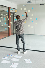 Rear view of African businessman standing in front of glass blackboard and making notes on adhesive...