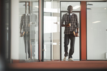 Fototapeta premium African young businessman in casual clothing walking along the office corridor and drinking coffee
