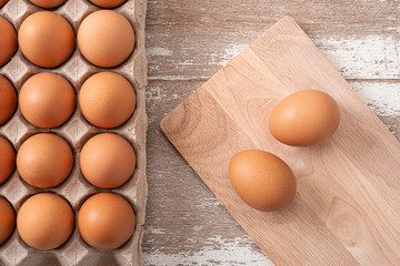 Two eggs on cutting board and many fresh raw brown chicken eggs in carton box on wooden rustic white table background. Top view.