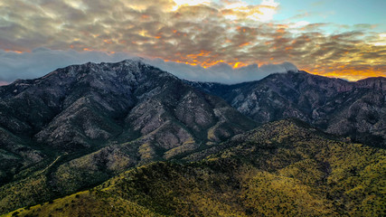 Aerial view of a sunset over a mountain range in Arizona/ The sky is orange and yellow and has many clouds while the mountains are dark and the landscape is green.