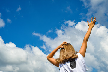 A woman stands with her back to the photographer in a white T-shirt and against a blue sky with clouds. Reaches his hand to the sky. Desire to move forward