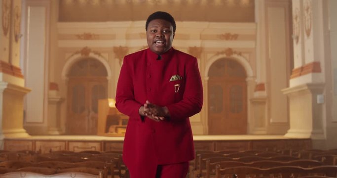 Afro american male performer in red suit looking to camera and singing while standing at row of wooden pews in hall. Smiling guy performing gospel music emotionally and dancing