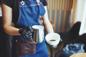 Female holds a milk jug and a cup of coffee, makes cappuccino, pouring whipped milk into espresso.Blurred image, selective focus