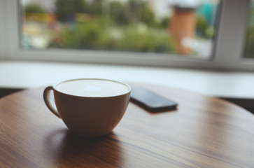 
Blurred image of cup of coffee latte and smart phone on table in cafe. Morning light falls from the window.Coffee break