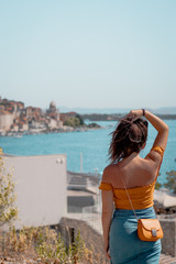 Back view of an attractive brunnete holding her long hair up, looking into the distance. Posing near the bright blue sea, outline of the town of Sibenik in Croatia in the distance