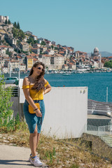 Attractive brunette wearing a blue skirt and an orange top posing outside near the sea on a warm summer day. Long hair flowing in the wind. Outline of Sibenik town in the distance. Influencer travels