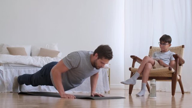 Family fitness at home, Father coach, Sports kid, Fun gymnastics. Boy is recording sports records while sitting on a chair at home. Man push up