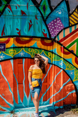 Fototapeta na wymiar Attractive brunette girl wearing a blue skirt and an orange top posing on a summer day with a colorful wall in the background. Yellow purse, stylish influencer travelling and modelling