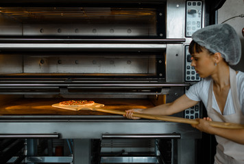 Young caucasian woman baker is holding a wood peel with fresh pizza and put it in an oven at a baking manufacture factory.