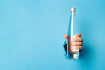 Man's hand holding a glass bottle with water on a bright blue background