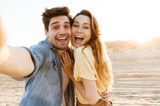 Image of young couple laughing and taking selfie photo on beach