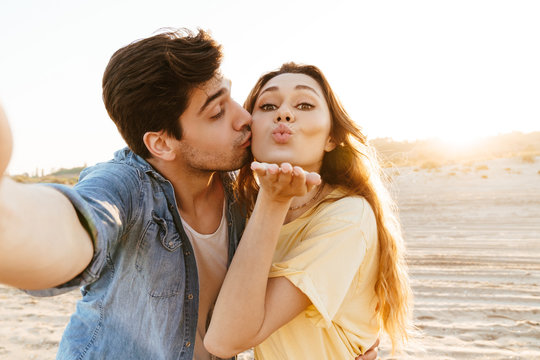Image of young couple kissing and taking selfie photo together on beach