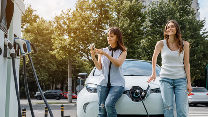 Two women's best friends are sitting on the electric car until one of them is using her smartphone to pay for charging her electric car.