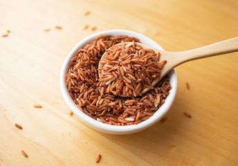 Closeup brown rice or red rice with wooden spoon and white ceramic bowl on wood table top. Healthy organic food concept.