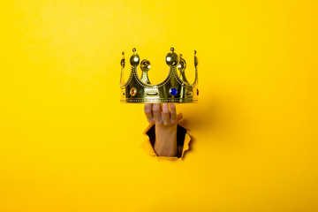 Female hand holds a gold crown on a bright yellow background. - obrazy, fototapety, plakaty