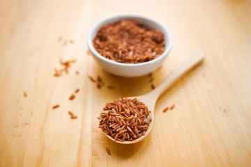 Closeup brown rice or red rice with wooden spoon and white ceramic bowl on wood table top. Healthy organic food concept.