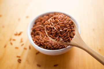 Closeup brown rice or red rice with wooden spoon and white ceramic bowl on wood table top. Healthy organic food concept.