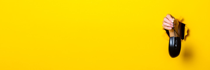 Female hand holding a black computer mouse on a bright yellow background. Banner.