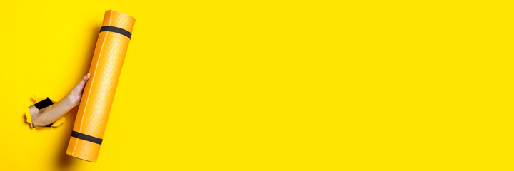Female hand holding an orange yoga mat on a bright yellow background. Banner