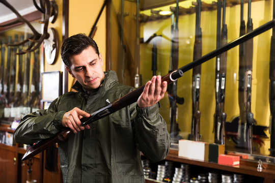 Portrait of man in hunting raincoat in gun store carefully selecting rifle