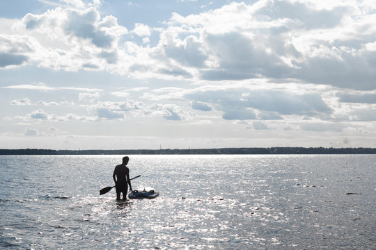 Silhouette of a man standing in water near SUP board with a paddle in hand. Learning to ride on a SUP board.