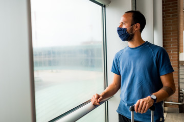 young man wearing protective face mask, traveling in the time of covid19 pandemic inside an airport, looking out of window with worried and upset expression. corona virus and travel industry