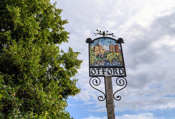 The Otford village sign in Otford, Kent, UK. The sign is on a grass area near the village pond. Images shows just the Otford sign and a tree.