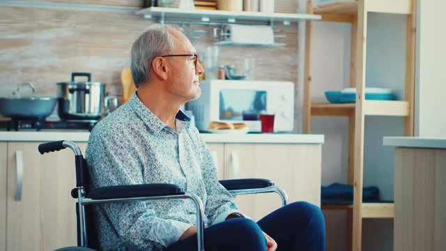 Lonely invalid man sitting in wheelchair looking through kitchen window. Elderly handicapped pensioner after injury and rehab, paralysis and disability for depressed invalid full of sorrow, worry and