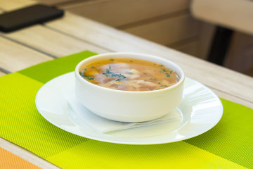 Noodle soup in a summer cafe. Soup with yellow broth in a white serving plate. Top view of the dish. Wooden white table in the cafe. Healthy chicken broth with meat and noodles. Lunch time.
