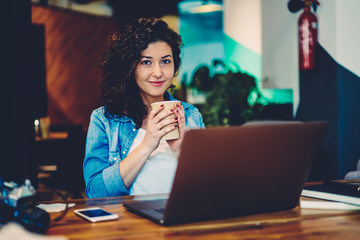 Half length portrait of beautiful female copywriter enjoying tasty beverage during working break in coffee shop.Gorgeous positive hipster girl looking at camera and holding take away drink indoors