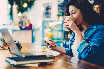 Young female person enjoying takeaway coffee while sharing news and chatting with followers via smartphone and internet connection.Cropped image of hipster girl drinking beverage and checking email